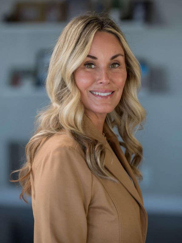 Confident woman with blonde hair wearing a beige blazer smiles warmly.