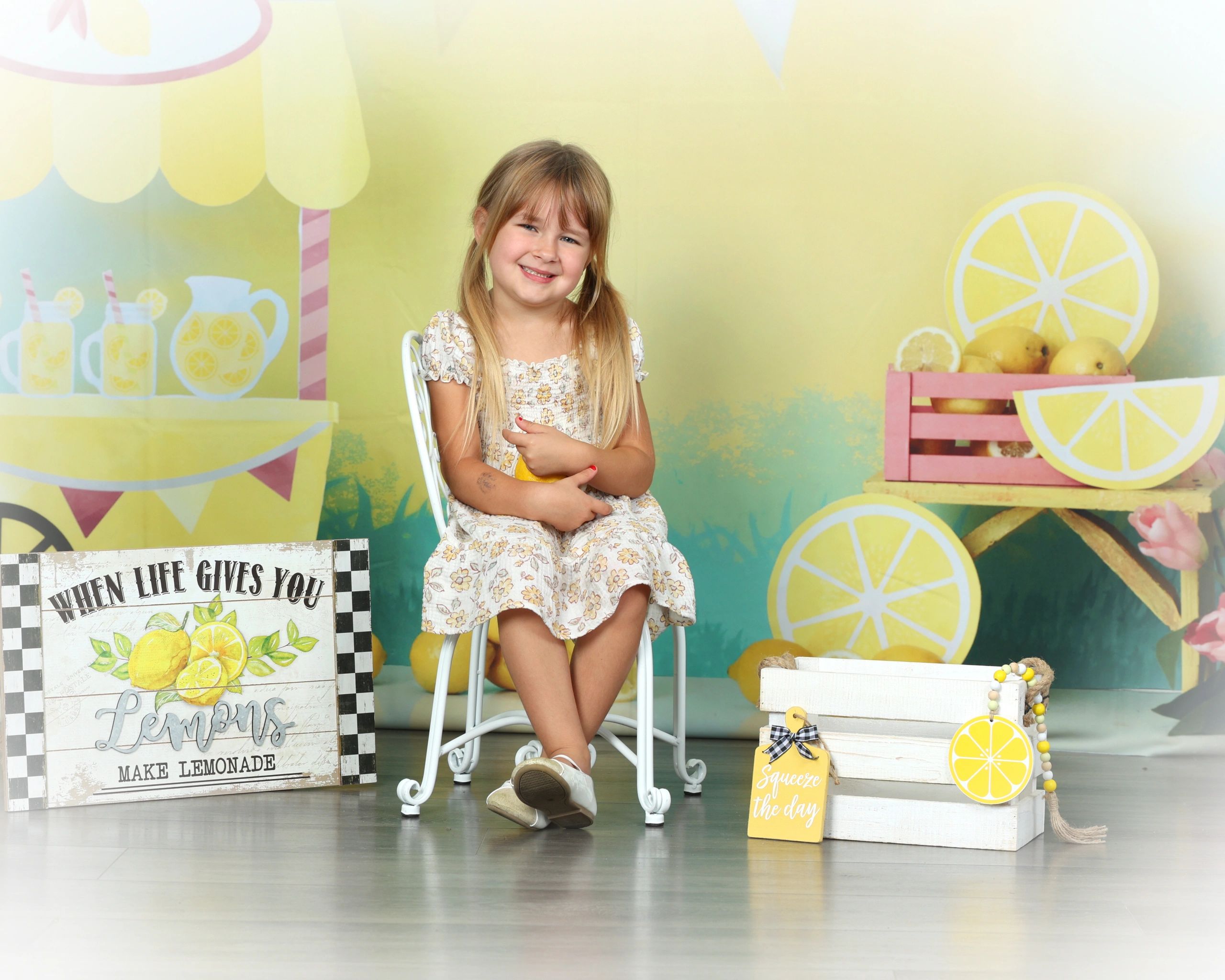 Young girl in yellow flower print dress sitting in white chair posing with lemonade backdrop