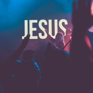 Crowd of worshipers singing along to band on stage with word in background that says "Jesus" in whit