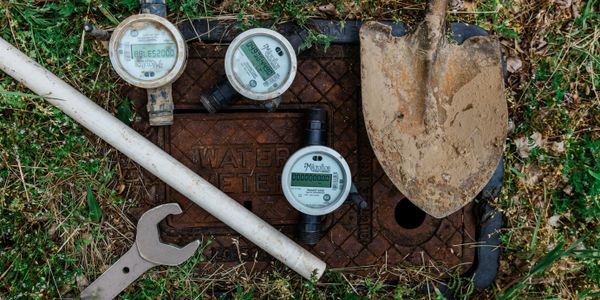 Water meters, a wrench, a pipe, and a dirty shovel on grass near a water meter cover.