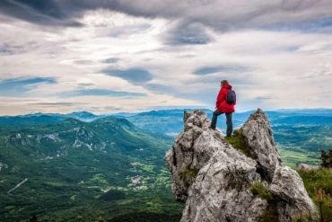 Person in red jacket on rocky cliff overlooking green mountains.