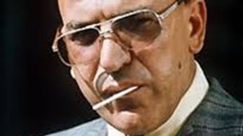 A bald man in a suit wearing sunglasses with a cigarette.