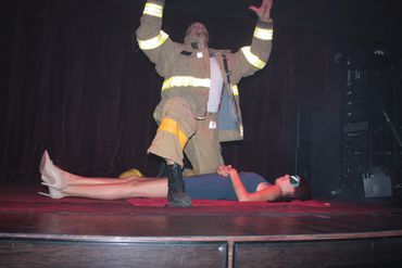 Jessy kneeling in a fireman outfit on stage at a strip club. Woman laying on her back underneath