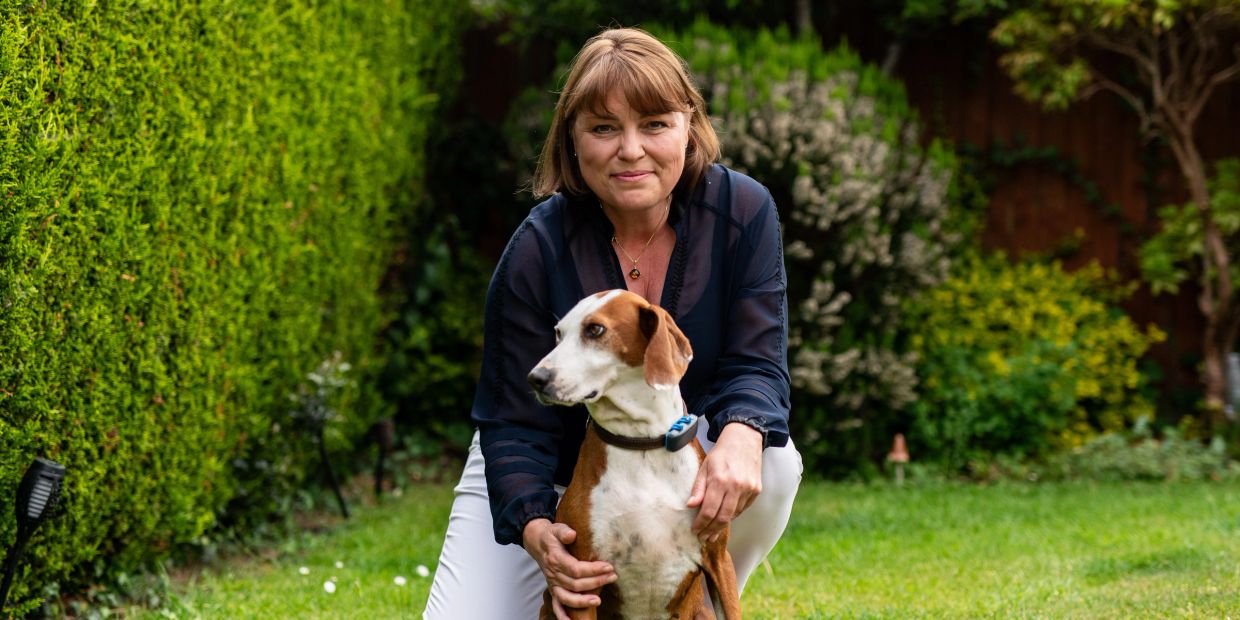 A woman kneels in a garden with a brown and white dog, surrounded by greenery.