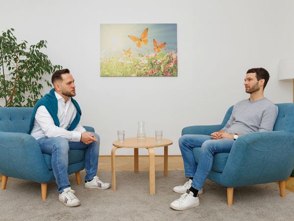 Two men engaged in a calm conversation in a cozy room with blue chairs.