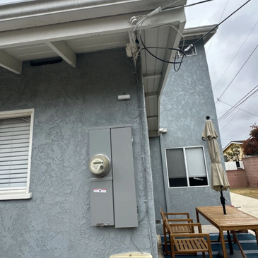 Outdoor electrical meter and patio furniture beside a gray textured house wall under a cloudy sky.