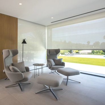 Modern lounge area with gray chairs and ottomans by a large window overlooking a garden.