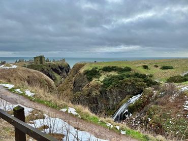 Ruined castle on cliffs by the ocean with a waterfall and snowy patches.