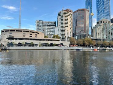 City skyline with buildings reflecting on a calm river under a clear sky.