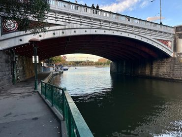 A scenic riverside view under a decorative bridge with people walking above and boats on the water.