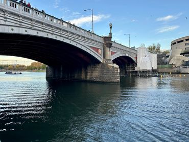 A metal and stone bridge over a calm river with people walking on it.