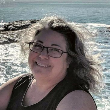 Smiling woman with grey hair and black glasses in front of water.
