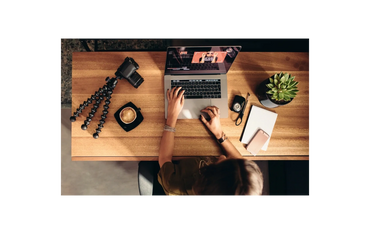 Person working on a laptop at a wooden desk with camera gear and coffee.