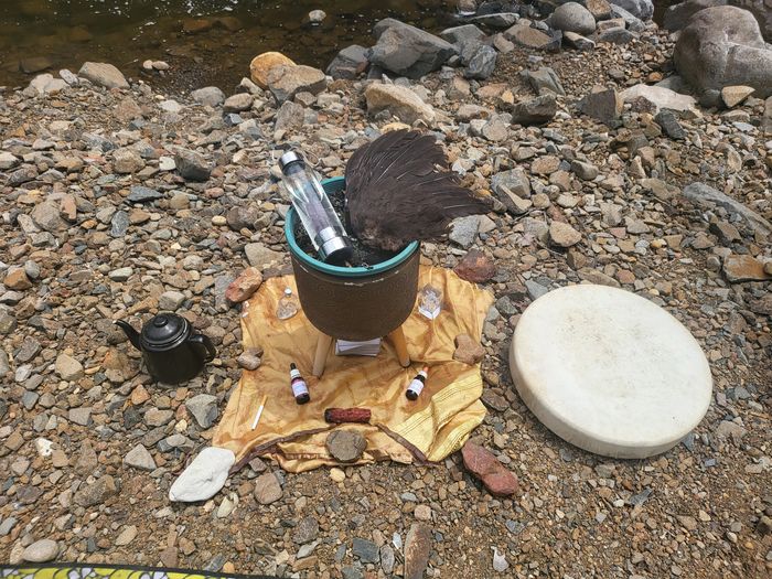 Outdoor ritual setup with feathers, bottles, and drum on rocky ground.