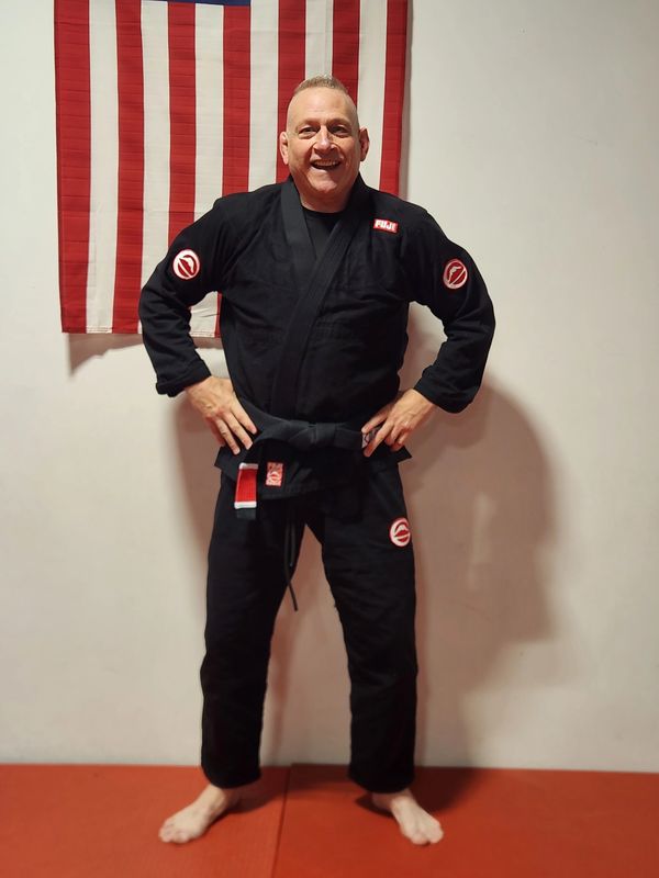 Man in black martial arts gi with black belt standing in front of American flag.