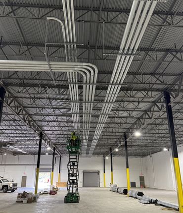 Worker on green lift installs metal conduits on warehouse ceiling.