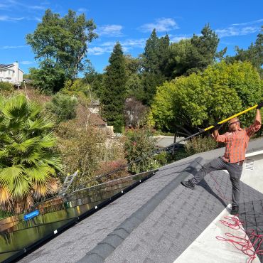 Man cleaning solar panels on a rooftop with a long pole.