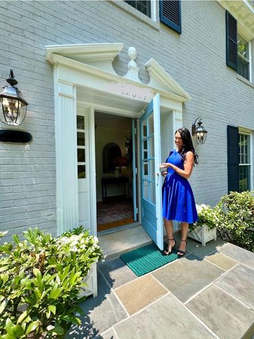 Woman in blue dress opens a charming front door of a brick house with greenery.