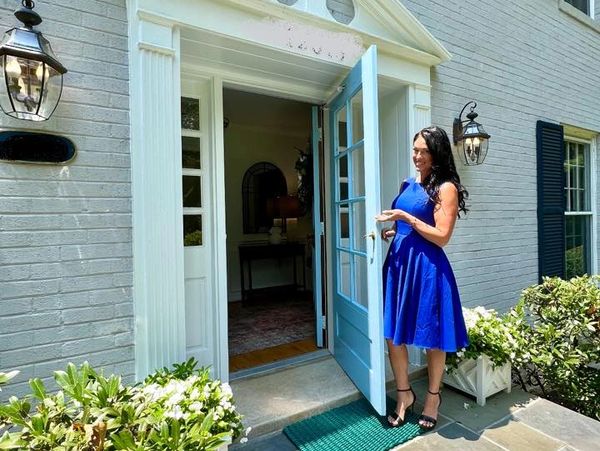 Woman in blue dress opens a charming front door of a brick house with greenery.