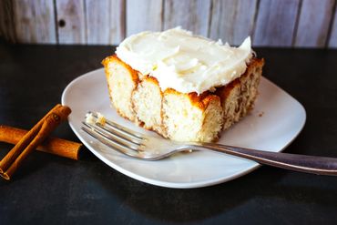 Slice of cinnamon roll cake with cream cheese frosting on a white plate.