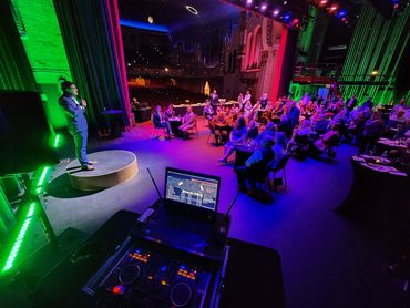A man in a blue suit speaks on stage to an audience in a colorfully lit venue.