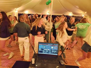 A lively group of young people dancing under a tent with DJ equipment in the foreground.
