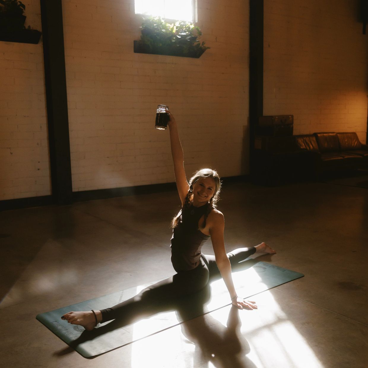 Woman doing splits on a yoga mat, holding a drink and smiling.