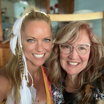 Two smiling women posing closely, one with feathers in her hair.