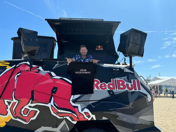 Man holding a Cream Entertainment shirt on a Red Bull vehicle at the beach.