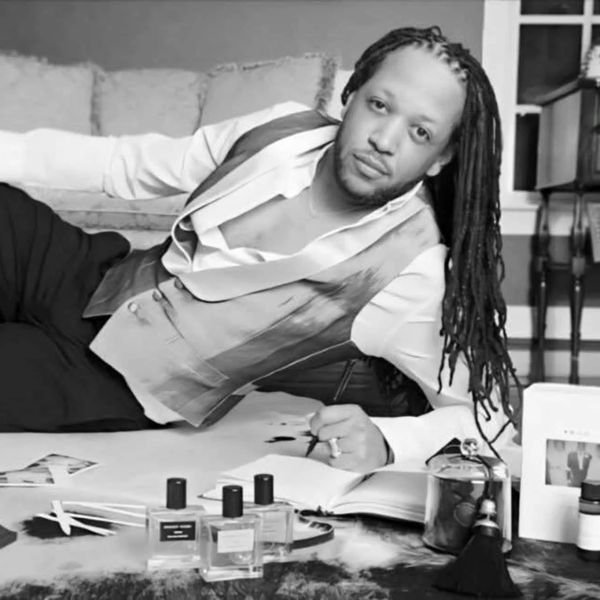 Man with dreadlocks lying on the floor surrounded by perfume bottles.
