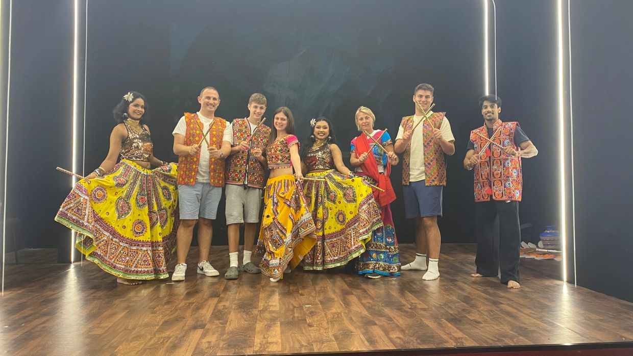 Group of people in traditional colorful Indian attire posing with sticks on a stage.