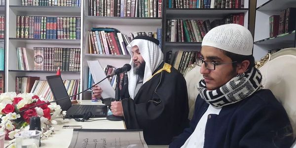 Two men in traditional attire reading and speaking in a library.