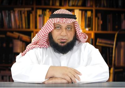Middle-aged man in traditional Saudi attire sitting at a desk with bookshelves behind.