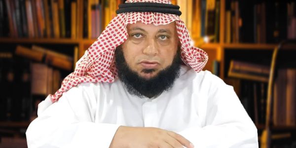 A man in traditional Middle Eastern attire sitting in front of bookshelves.