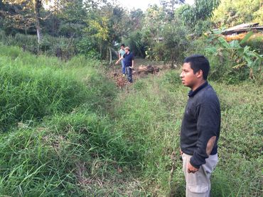 Three men stand in a grassy, wooded area, seemingly inspecting something on the ground.