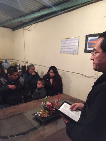 Man reading from a tablet to a small group in a simple room with candles on the table.