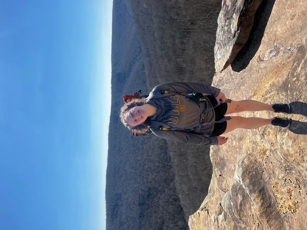 Smiling woman hiking on a rocky cliff with forested hills in the background.