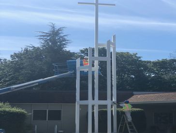 Workers refurbishing a tall white church cross structure on a sunny day.