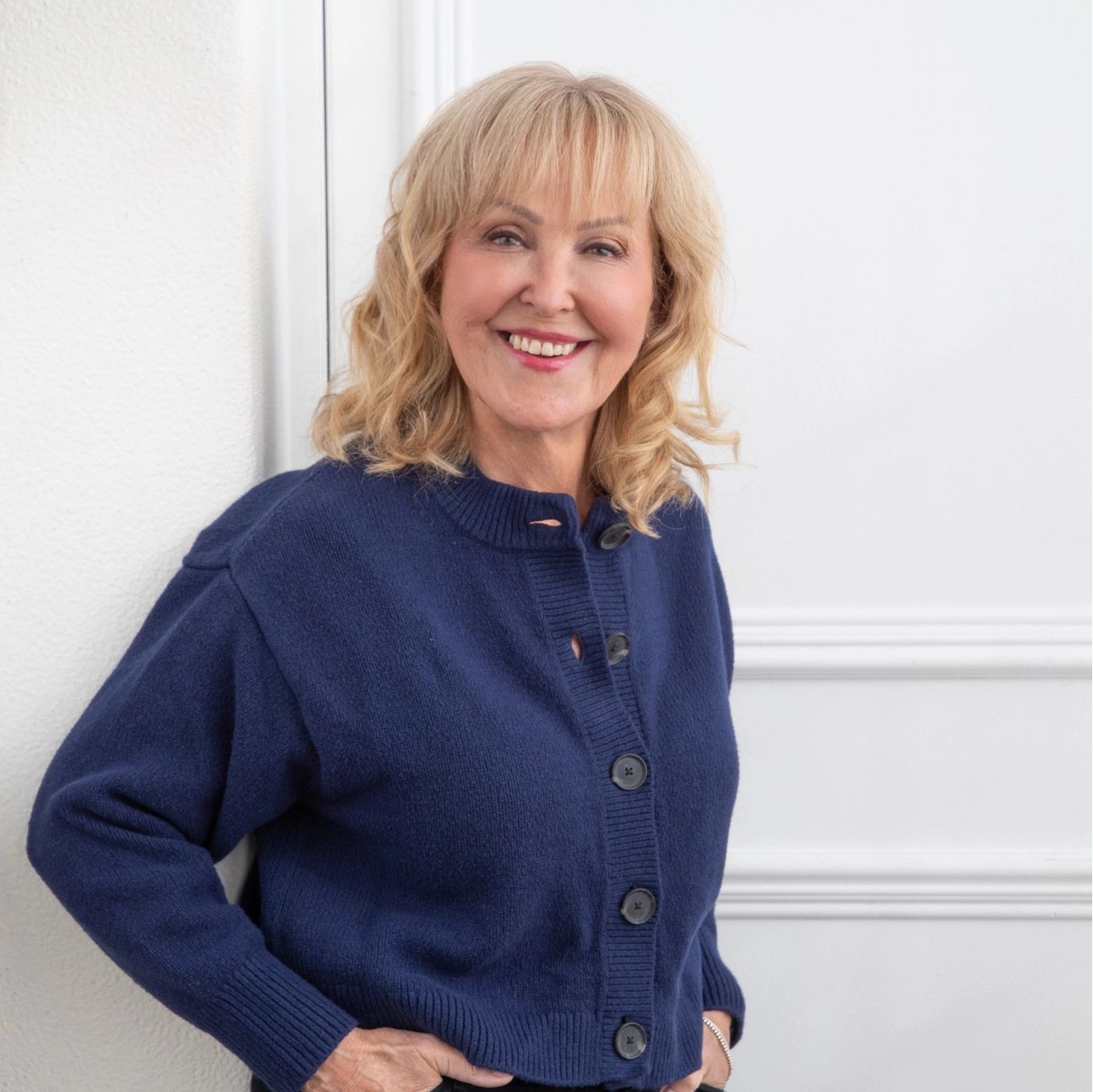 Smiling older woman in a navy blue cardigan standing against a white wall.