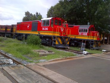 Two bright red and yellow diesel locomotives on tracks near greenery and a platform.