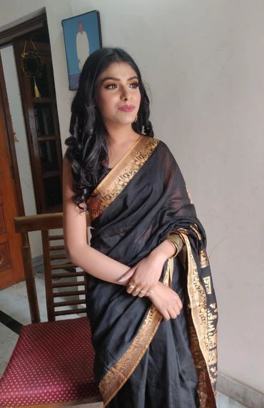A woman wearing a black saree with golden border, standing indoors.