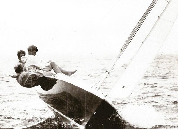 Three people leaning out of a small sailboat on choppy water.