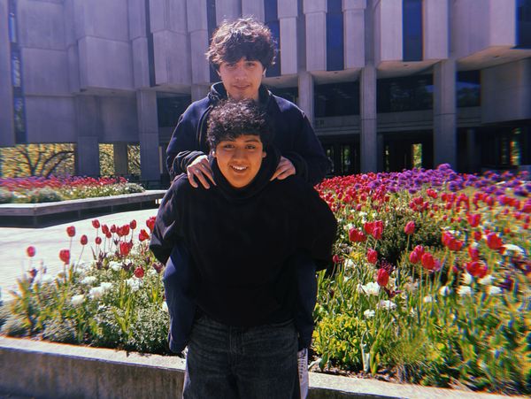 Two young men posing playfully in front of a colorful flower garden outdoors.