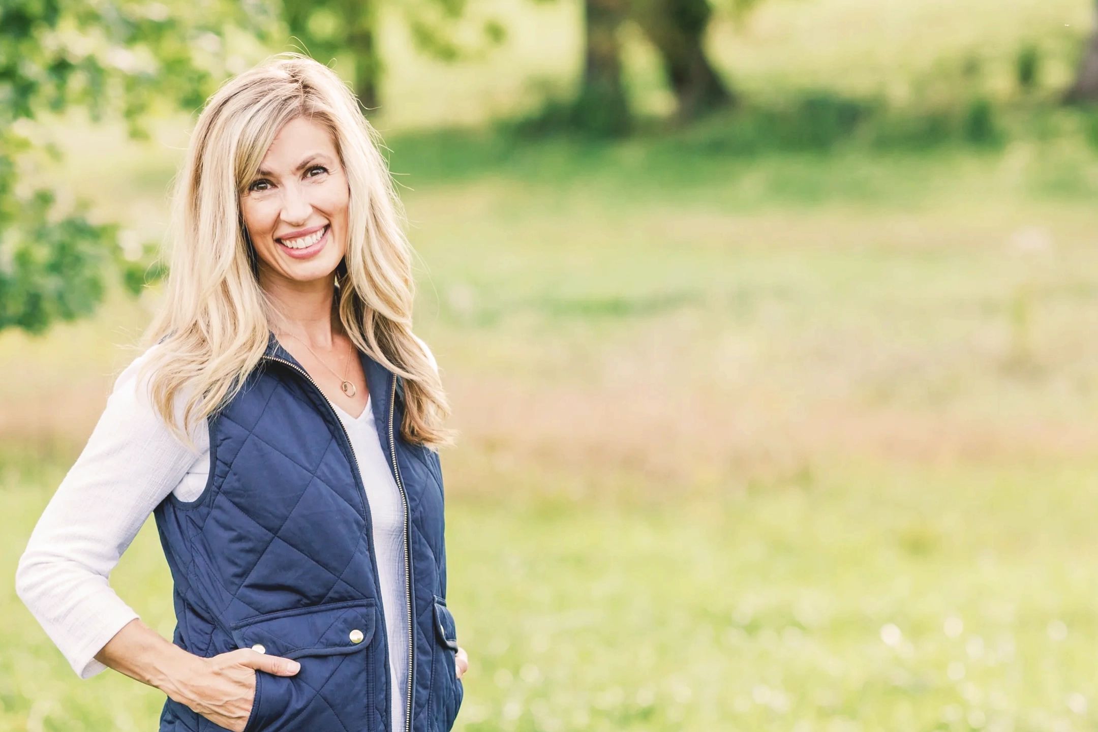 Smiling woman in a navy vest outdoors with green foliage background.