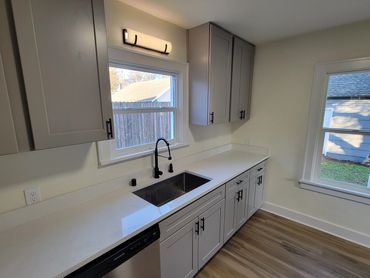 Modern kitchen with gray cabinets, white countertops, and a black sink faucet under a window.