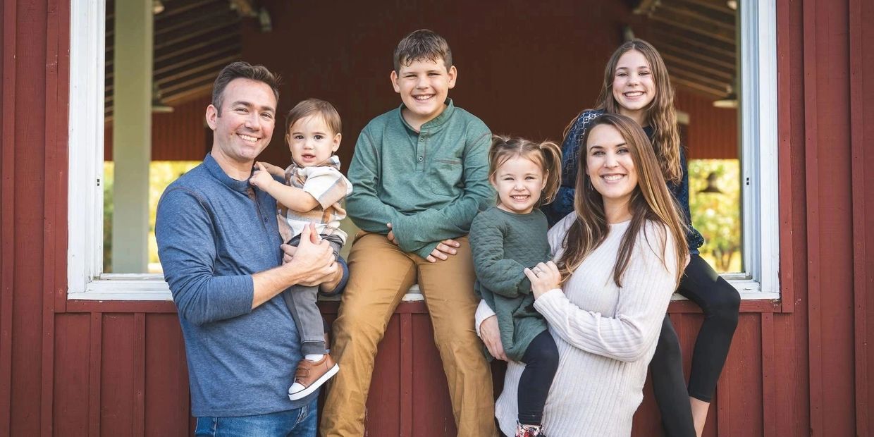 A happy family of six posing together outside a red wooden building.