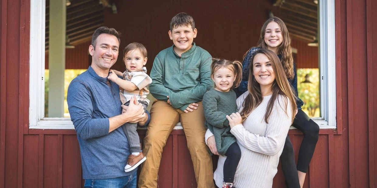 A happy family of six posing together outside a red wooden building.