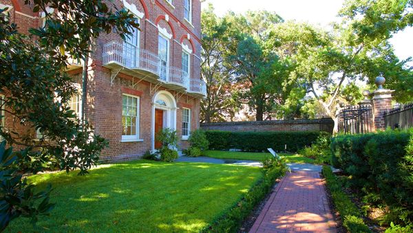 Historic brick building with a lush garden and tree-lined path.
