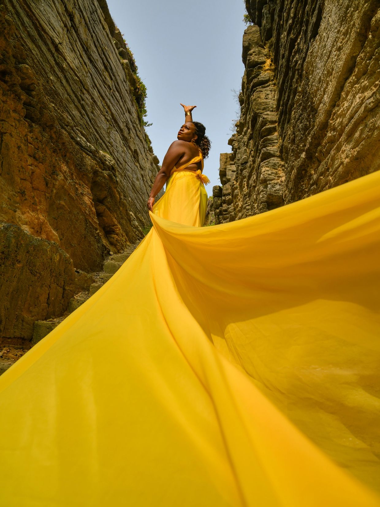 Woman in a flowing yellow dress between rock walls, reaching upward.