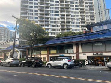 Street view with parked cars, a woman walking, and a building with shops and a tall apartment in the background.